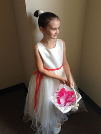 Young flower girl holding basket looking out window while standing against wall