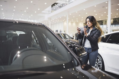 Young woman photographing new car through mobile phone at showroom