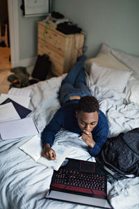 High angle view of teenage boy reading book while using laptop on bed at home