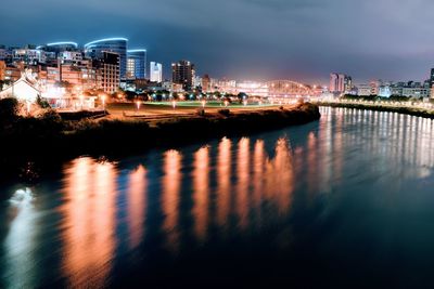 Illuminated buildings by river against sky at night