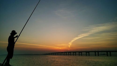 Silhouette man fishing in sea against sky during sunset