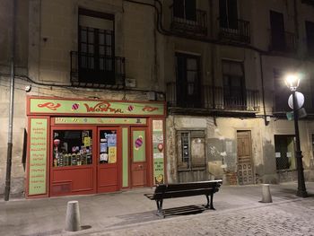 Empty bench on illuminated street by building at night