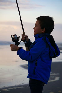 Side view of holding fishing rod standing on beach during sunset