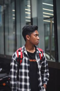 Young man looking away while standing against window