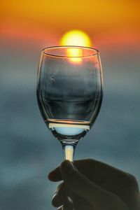 Close-up of hand holding wineglass against sky during sunset