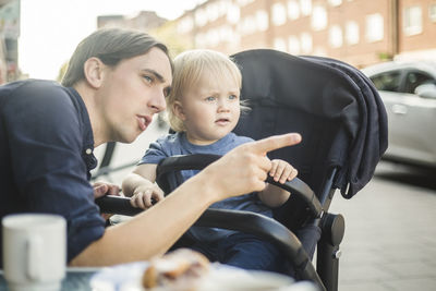 Father pointing while looking away with son at sidewalk cafe in city