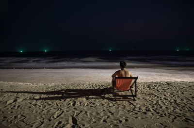 Rear view of man sitting on chair at beach during night