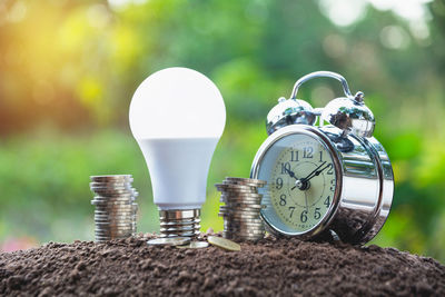 Close-up of alarm clock by coins stack and led bulb on mud