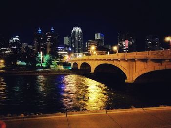 Bridge over river by illuminated buildings against sky at night