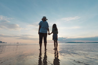 Rear view of mother and daughter holding hands while standing at beach against sky