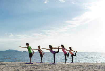 Young friends practicing yoga in warrior position on shore at beach during sunny day