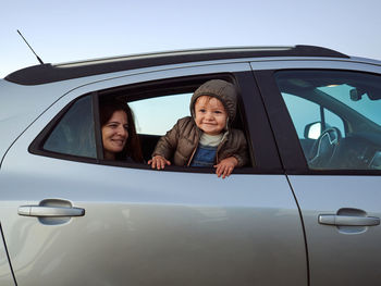Cheerful cute toddler peering out of car window while sitting on back seat with mother and looking away