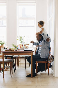 Father carrying daughter on shoulder while using laptop at table in house