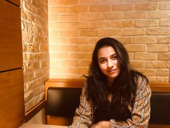 Portrait of smiling young woman sitting on seat against brick wall