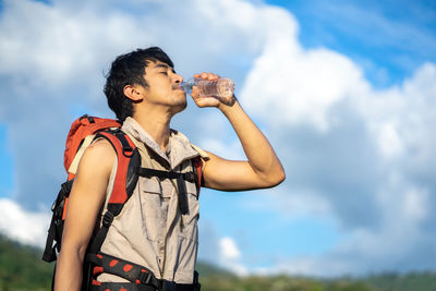 Side view of young man drinking water while standing against sky