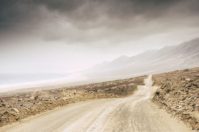 Empty dirt road leading towards mountains at desert against sky