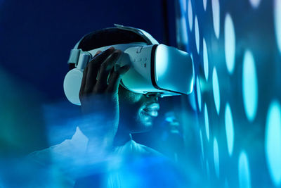 Side view of concentrated young african american in white t shirt and modern vr goggles interacting with virtual object in dark studio with neon illumination