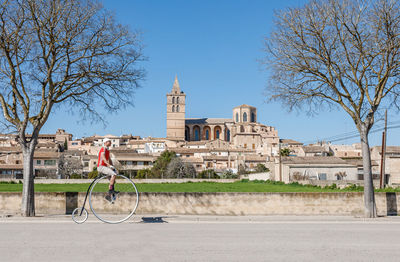 From below side view of unrecognizable male in casual clothes riding retro high wheel bike along street against facades of small buildings and ancient palace at horizon in downtown under cloudless blue sky in sunny day