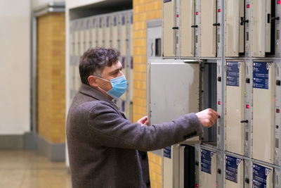 Male traveler wearing a protective surgical mask opening or closing a train station locker cabinet