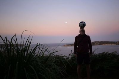 Man balancing soccer ball on head against sea during sunset