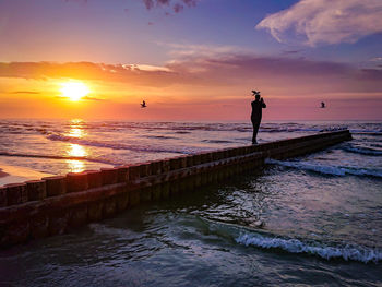 Silhouette man standing on wooden post amidst sea against sky during sunset