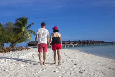 Rear view of couple walking on shore at beach against blue sky