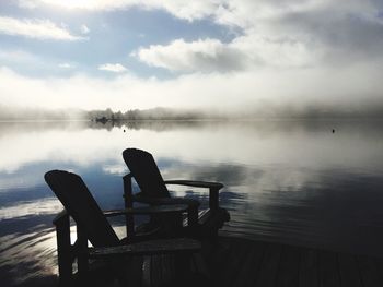 Empty chairs on boardwalk by lake against cloudy sky