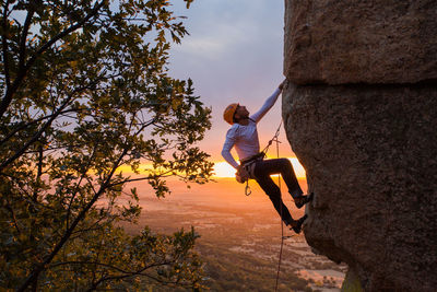 Side view of active climber in protective helmet hanging on mountain holding on to slope in warm light of sunset