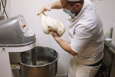 Mid adult chef preparing dough for bread by electric mixer in bakery during covid-19