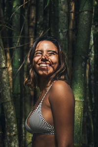 Side view of content young female traveler in swimwear with shadow on face looking away against bamboo twigs