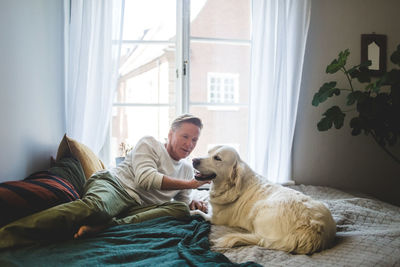 Senior man stroking dog while leaning on bed at home