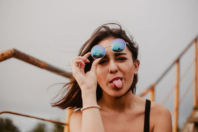 Young woman in sunglasses sticking out tongue against sky