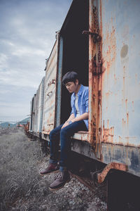 Sad young man sitting on abandoned freight train against sky