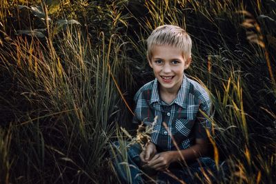 High angle portrait of smiling boy sitting on field amidst plants