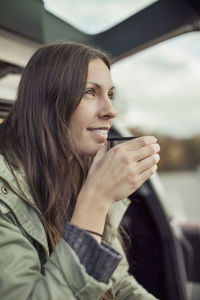 Young woman looking away while drinking coffee at car's trunk