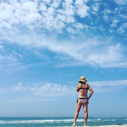 Rear view of woman wearing bikini standing on sea shore at beach against sky