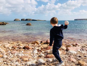 Rear view of boy throwing pebble stones at beach against sky