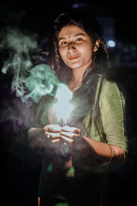 Portrait of beautiful young woman holding illuminated sparkler while standing at night