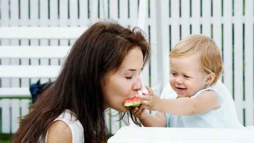 Summer, in the garden, a funny one-year-old blonde girl treats her mother with watermelon, feeds