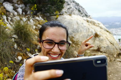 Smiling woman gesturing while taking selfie against rock