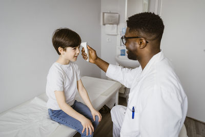 Young male pediatrician measuring temperature of boy using infrared thermometer at clinic