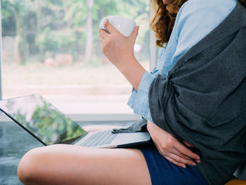 Midsection of woman holding coffee cup while sitting with laptop