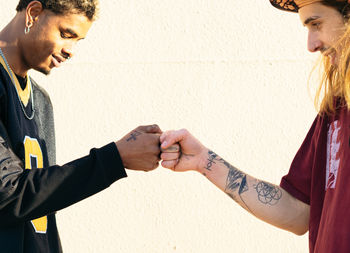 Side view of crop young multiracial hipster men in cool outfit with piercings and tattoos greeting each other on light background