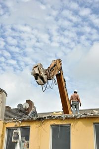 Low angle view of man standing on roof by crane against cloudy sky