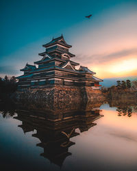 Traditional building by lake against sky during sunset