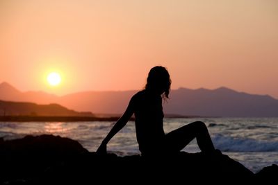 Silhouette woman sitting on rock at beach against clear sky during sunset