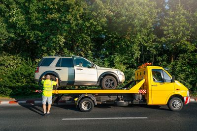 Tow truck and broken car on road against trees in city