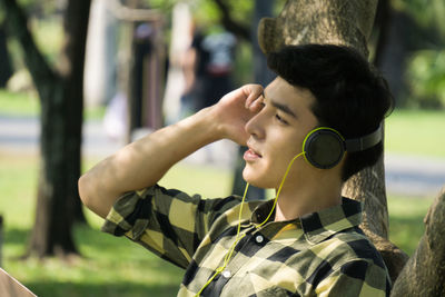 Side view of young man wearing headphones by tree trunk at park