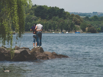 Rear view of friends standing on rock at lakeshore against sky