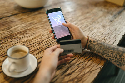 From above of crop unrecognizable male with plastic card making purchase for order and using mobile application for online shopping on smartphone while sitting at table in cafe